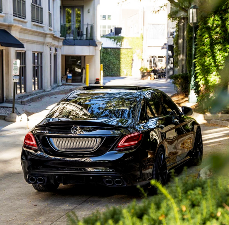 A black Mercedes-Benz C43 car is parked on a narrow, sunlit street lined with buildings and greenery. A sign reads "2 HOUR PARKING CUSTOMER PARKING ONLY."