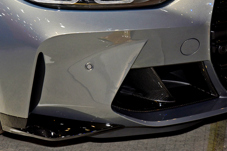 A sleek, silver car's front bumper features aerodynamic vents and textured black details, situated in a well-lit indoor setting.