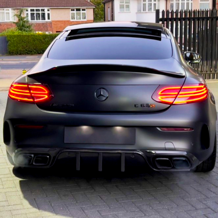 A black Mercedes-Benz C 63 S AMG is parked, with illuminated tail lights, on a paved driveway. Houses and a black metal fence are visible in the background.