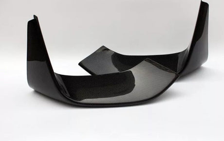 Two carbon fiber car parts, possibly rear exhaust diffusers, are positioned diagonally on a solid white surface, highlighting their glossy black finish and textured pattern.