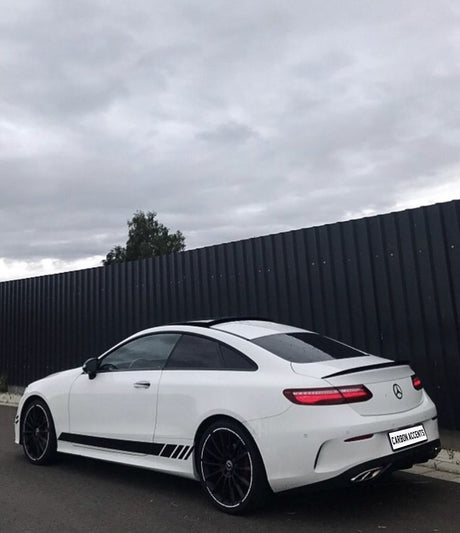 A white sports coupe is parked on a street beside a tall, dark fence under a cloudy sky. The license plate reads "CARBON ACCENTS."