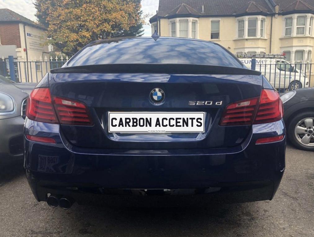 A blue BMW 520d is parked with its rear facing the camera. The license plate reads "CARBON ACCENTS." Nearby are other cars, buildings, and a tree.