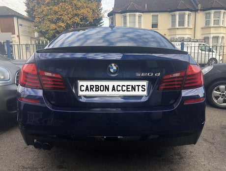 A blue BMW 520d is parked with its rear facing the camera. The license plate reads "CARBON ACCENTS." Nearby are other cars, buildings, and a tree.