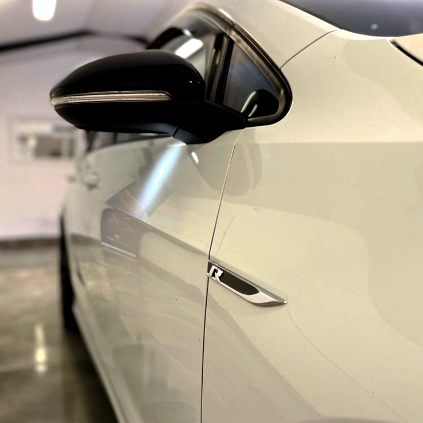 A close-up of a white car's side, featuring a black side mirror and an "R" badge, set in a brightly lit indoor space with polished floors.