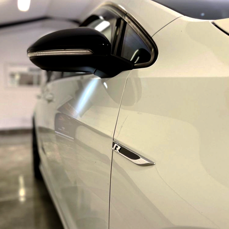 A close-up of a white car's side, featuring a black side mirror and an "R" badge, set in a brightly lit indoor space with polished floors.
