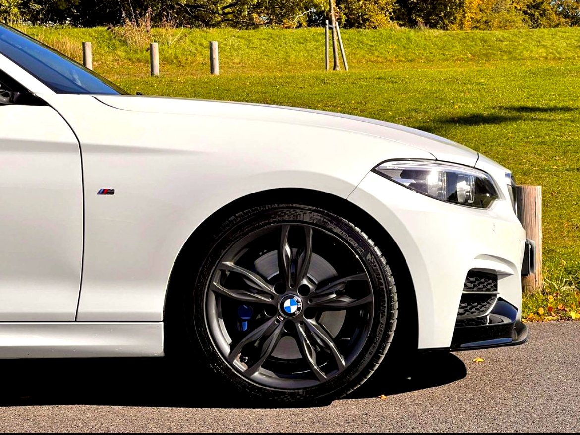 A white car is parked on a paved road. Its front wheel is turned slightly, showcasing a five-spoke alloy rim. The background features a grassy hill and wooden posts.