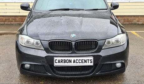 A black BMW car with water droplets on the hood is parked. The license plate reads "CARBON ACCENTS." The background features a brick wall and pavement.
