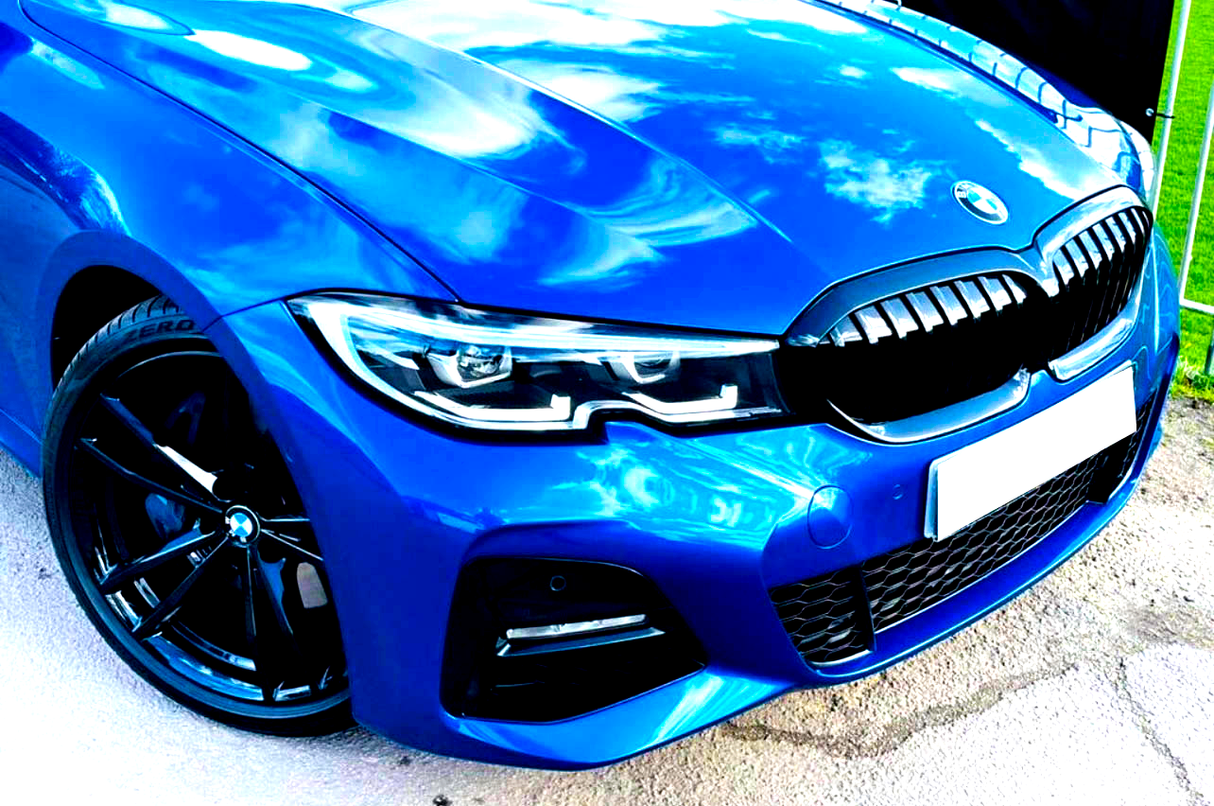 A blue car with a shiny exterior reflects clouds, highlighting a sleek headlight and distinct grille, parked on a textured surface beside green grass.
