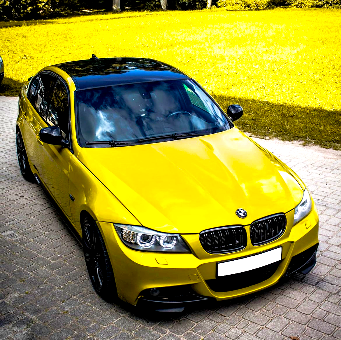 A yellow car is parked on a paved driveway. It features a glossy black roof and is surrounded by a grassy area with scattered trees in the background.
