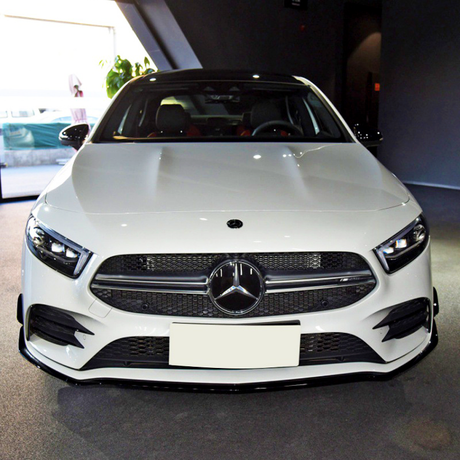A white Mercedes-Benz car is parked indoors on a carpeted floor, with natural light coming from a left-side window, highlighting its front grille and logo.