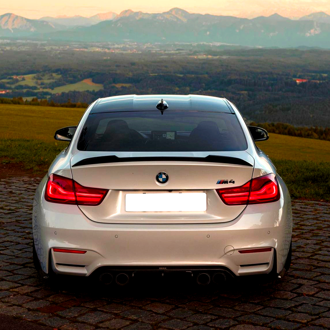 A silver BMW M4 is parked on a cobblestone path, rear view facing the camera, with a scenic backdrop of green fields and distant mountains under a clear sky.