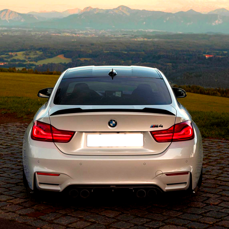 A silver BMW M4 is parked on a cobblestone path, rear view facing the camera, with a scenic backdrop of green fields and distant mountains under a clear sky.