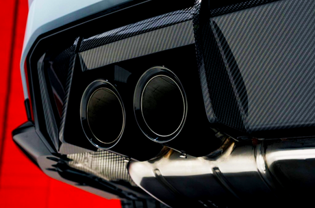 Twin exhaust pipes, metallic and polished, positioned at the rear of a vehicle with carbon fiber trim, against a bright red backdrop, highlighting the sleek automotive design.