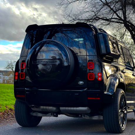 A black vehicle, with the word "DEFENDER" on the spare tire cover, is parked on a paved road. It's surrounded by trees and a grassy area under a cloudy sky.