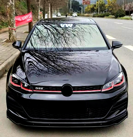 A shiny black car with red accents is parked on a tree-lined street. The windshield displays the letters "VW," and tree reflections are visible on the hood.