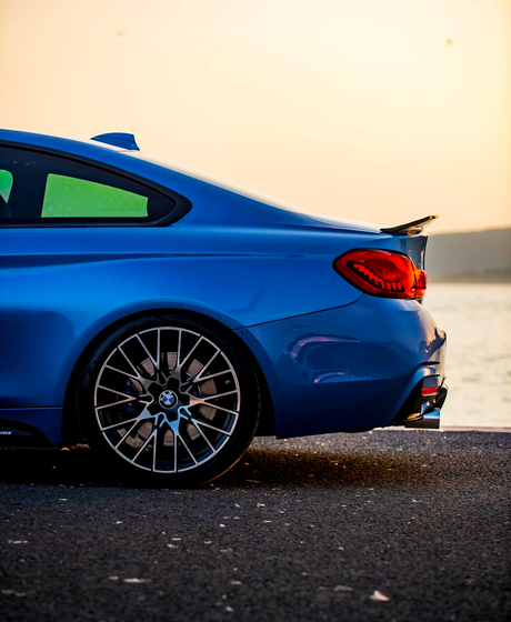 A blue sports car parked by a waterfront, showcasing its rear section with distinctive taillights and a prominent wheel in the foreground, against a serene sunset backdrop.