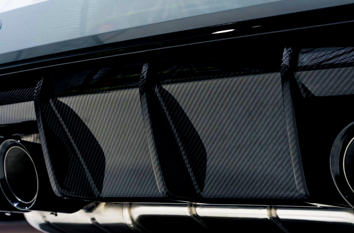 Dual exhaust pipes protrude from a sleek car rear diffuser with a carbon fiber texture, emphasizing performance and aerodynamics. The setting appears to be a modern garage or showroom.