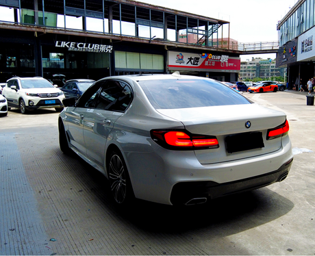 A silver BMW sedan is parked in a bustling urban setting, surrounded by other cars near buildings. Signs read "LIKE CLUB," "MO CLUB," and more in the background.