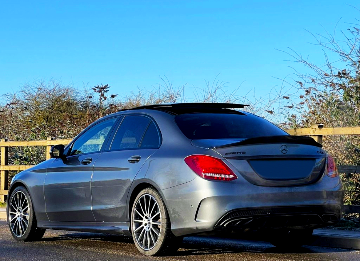 A gray Mercedes AMG car is parked on a road beside a wooden fence. The sky is clear, and tall shrubs line the background.