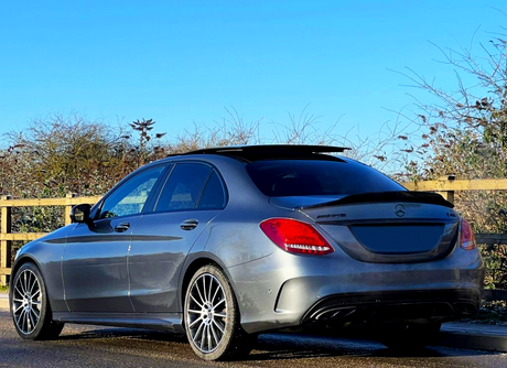 A gray Mercedes AMG car is parked on a road beside a wooden fence. The sky is clear, and tall shrubs line the background.