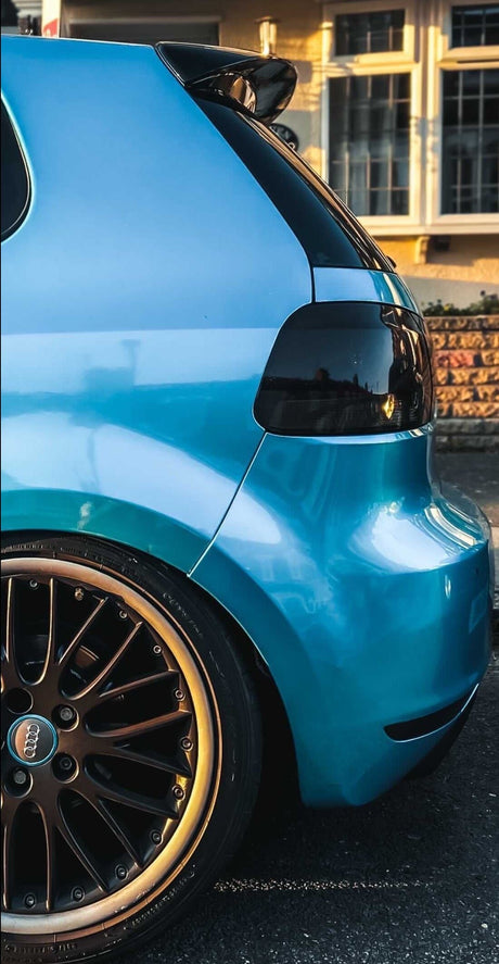 A blue car's rear end, showcasing tinted taillights and bronzed alloy wheels, is parked on a street. The background includes a building with warm-colored windows reflecting sunlight.