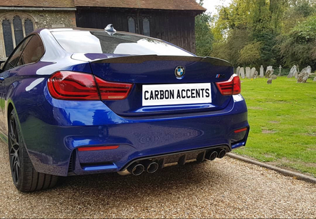 A blue BMW with the license plate "CARBON ACCENTS" is parked on a gravel path near a historic church building, surrounded by lush greenery and a small graveyard.
