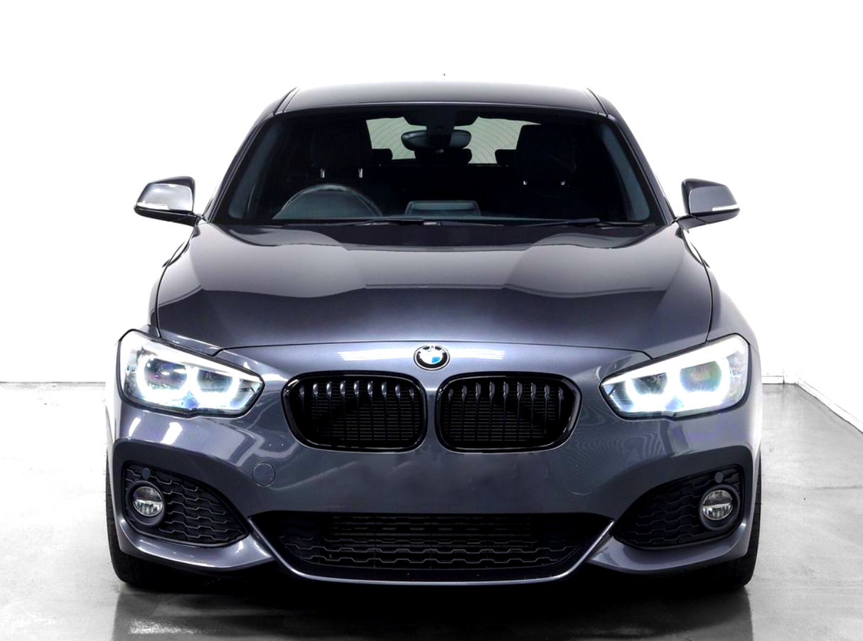 A gray BMW car, featuring illuminated headlights, is stationary in a bright, plain white environment, highlighting its sleek design and curved contours.