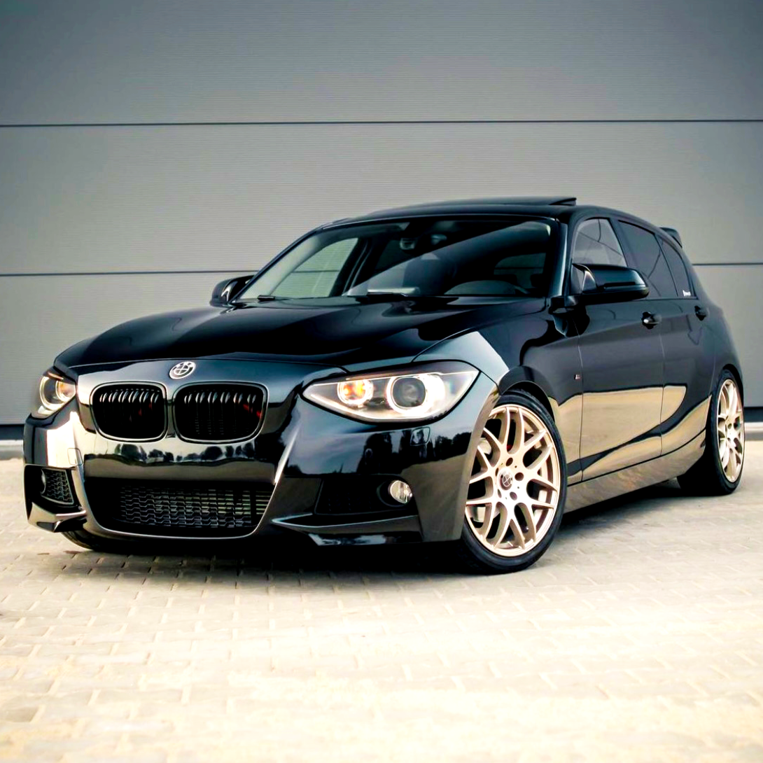 A sleek black car with a sporty design is parked on a paved surface in front of a plain gray wall, featuring shiny alloy wheels and prominent front headlights.