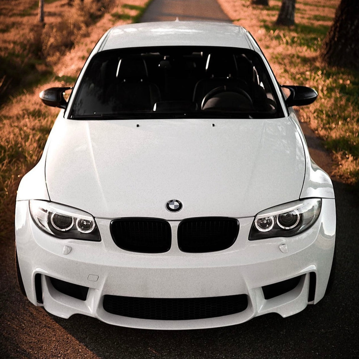 A white BMW car is parked on a rural road with grassy fields on either side, under a warm evening light.