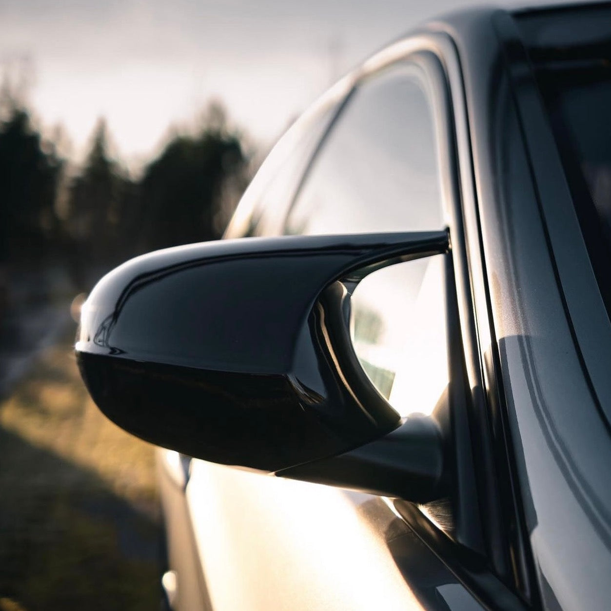 Car side mirror reflects sunlight, with a sleek black finish. The vehicle is parked outdoors, and the background features blurred silhouettes of trees under a bright sky.