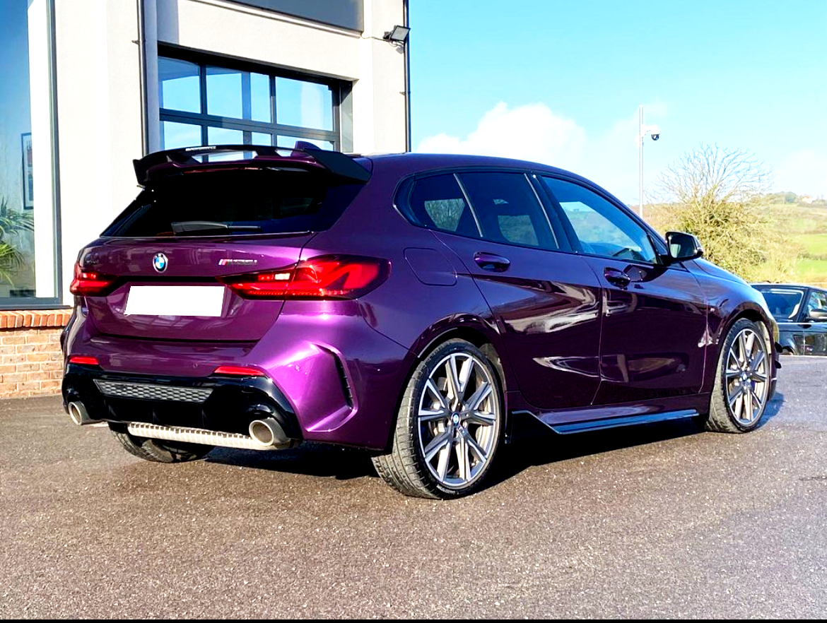 A purple BMW car is parked on a sunlit driveway beside a modern building with large windows. The surrounding landscape features green fields and a clear blue sky.