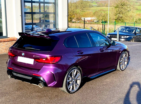 A purple hatchback car is parked on a paved surface beside a building with glass doors, with a scenic countryside landscape in the background.