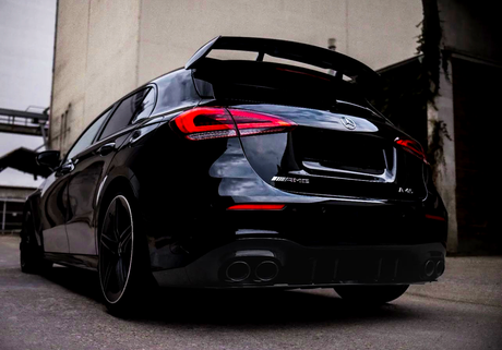 A black Mercedes-Benz AMG A-Class car is parked in an urban alley. It's viewed from the rear, emphasizing the distinctive taillights, spoiler, and dual exhausts. Text: "AMG" and "A45".