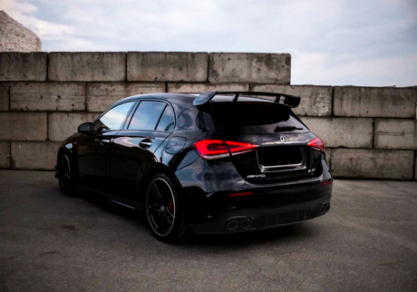 A sleek black hatchback car is parked, featuring a large rear spoiler. It is positioned in front of a concrete wall under a cloudy sky. Text on the car reads "AMG" and "A 45."