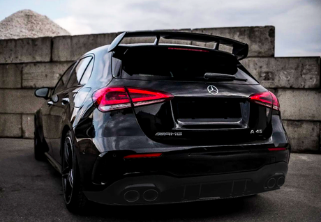 A black Mercedes A45 AMG with visible rear spoilers and dual exhausts is parked on a concrete surface, surrounded by high stone walls under a cloudy sky.