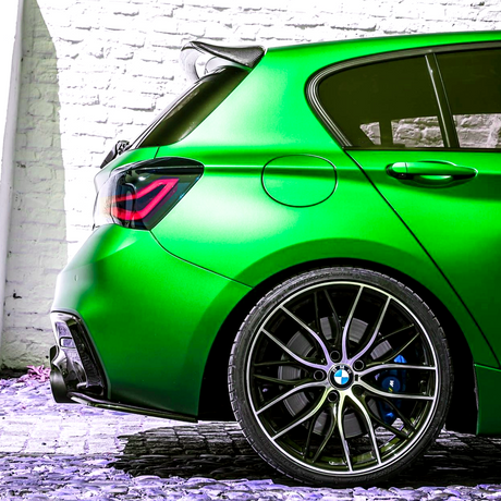 A vivid green car, positioned stationary, displays its rear wheel and taillight design against a white brick wall backdrop, emphasizing its sleek, sporty appearance.