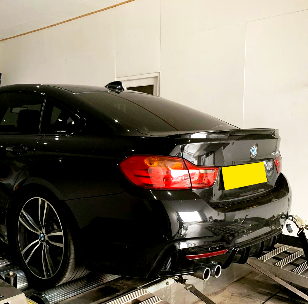A black car is secured on a testing rig in a white-walled room. The car has red tail lights illuminated and dual exhaust pipes. A strap is attached to its rear.