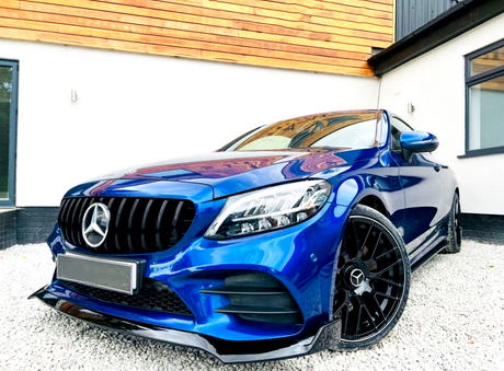 A blue Mercedes-Benz car is parked on gravel, featuring glossy black wheels and a prominent front grille, situated in front of a modern building with white walls and wooden accents.