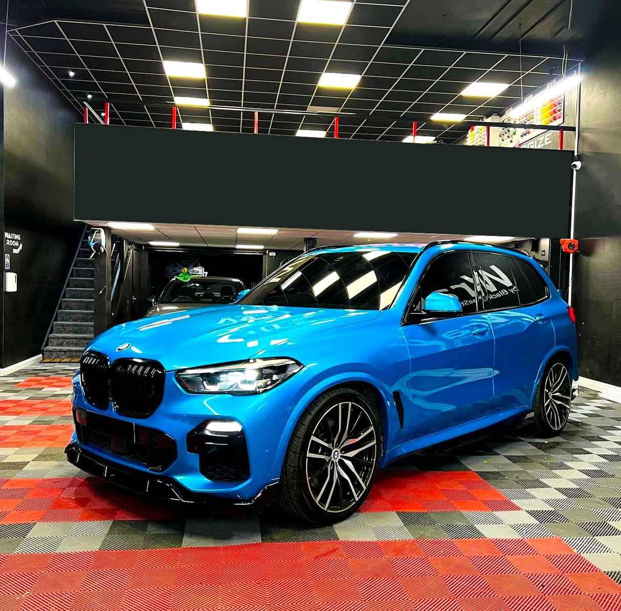 A blue BMW SUV is parked in a garage with a black ceiling and checkered red and black floor. "NAV" is written on the driver's side window.