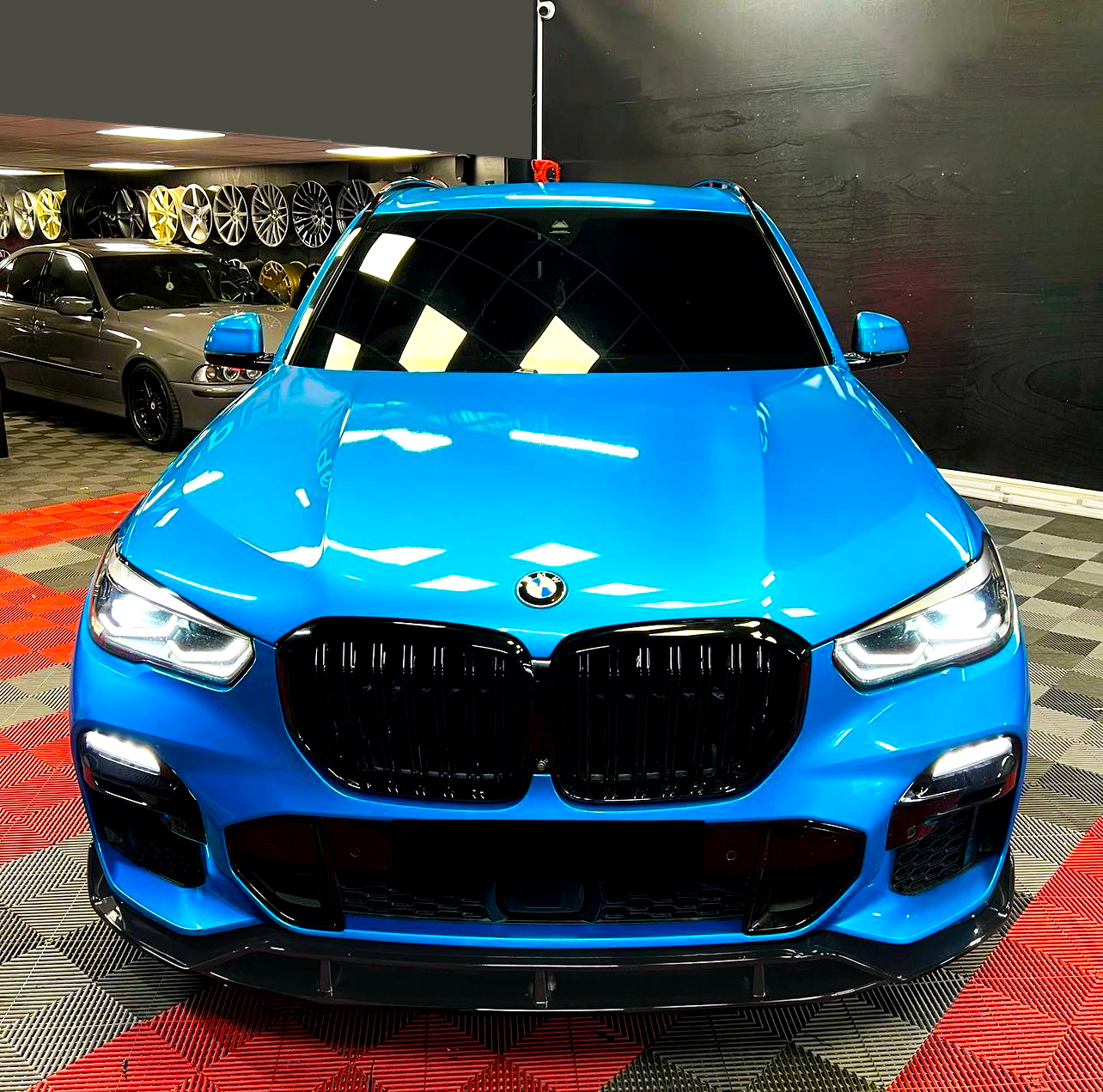 A blue BMW SUV is parked with headlights on in a showroom, surrounded by various wheels displayed on the wall and a checkered floor pattern.