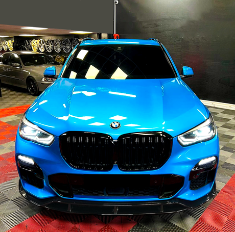 A blue BMW SUV is parked with headlights on in a showroom, surrounded by various wheels displayed on the wall and a checkered floor pattern.