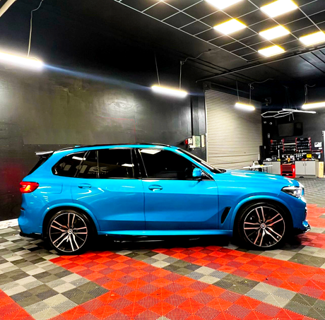 A bright blue SUV is parked in a garage with black walls and a checkered floor, under fluorescent lights, near a rolling door and shelves with tools.