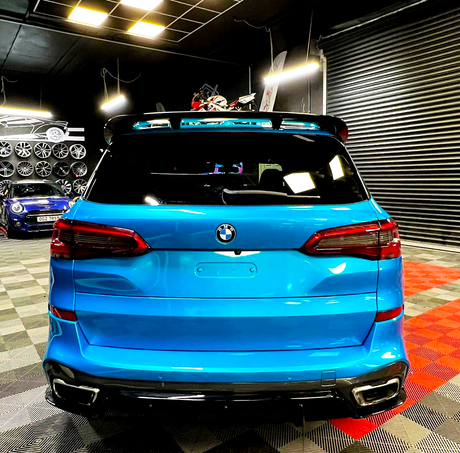 A bright blue BMW SUV is stationary in a garage with shelves of wheels and vehicles, like a blue sports car. The floor features checkered patterns, and lighting is overhead.
