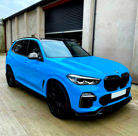 Bright blue SUV parked on a concrete surface, facing nearly forward. The car is in front of a tall, gray industrial building with a closed, dark roller door.