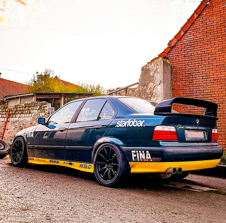 A modified blue car with a large rear spoiler and black wheels is parked on a street beside brick and stone walls. Text on the car reads "starobar," "FINA," "Michelin," and "HSD."