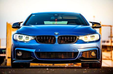A blue car with illuminated headlights is parked on a road. The background features a metal fence and a clear sky, highlighting the car's sleek design and front grille.