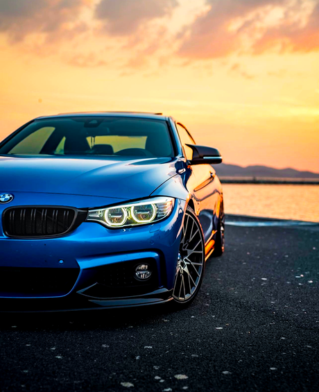 A blue sports car is parked on a paved surface, with its headlights on. The background features a vibrant sunset over a body of water and distant hills.