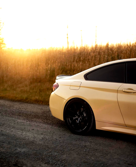 A yellow car is parked on a gravel road, with golden sunlight illuminating a field of tall grass in the background. The car's back wheel and tinted windows are visible.