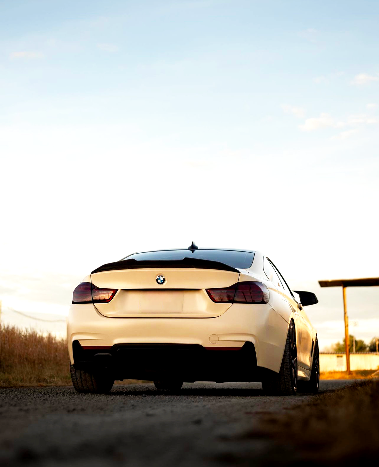 A white BMW car is parked on a dirt road during sunset, surrounded by open fields and a clear sky with a wooden structure in the background.