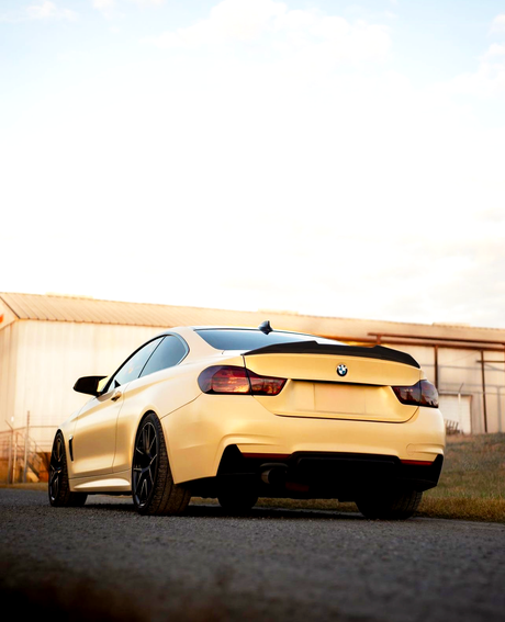 A yellow sports car is parked on a paved surface near a large, light-colored industrial building under a partly cloudy sky.